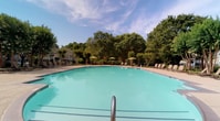 Located at 3700 W Steeplechase Way in Williamsburg, Virginia, this single-level community amenity space offers residents a well-appointed outdoor retreat and inviting interior common areas. The centerpiece of the outdoor space is a generously sized resort-style swimming pool with crystal-clear turquoise water, framed by a broad, tiled sundeck. The deck is furnished with rows of lounge chairs and shaded seating areas complete with yellow umbrellas, creating an ideal setting for relaxation. Mature trees and lush landscaping surround the pool area, providing a natural backdrop and a sense of seclusion. The multi-story residential buildings visible in the background, clad in neutral siding with balconies, lend a cohesive architectural character to the community. Inside, the common area entry corridor features warm, neutral-toned walls and durable slate-style tile flooring in earthy tones. Glass-panel entry doors allow natural light to flow through the hallway, which is accented with nautical-themed framed artwork, reinforcing a relaxed, coastal-inspired aesthetic. Safety features such as clearly marked fire alarm systems are present throughout. The overall ambiance of the space is casual, comfortable, and community-oriented.