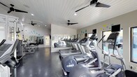 This single-level fitness center offers a bright and functional workout environment designed to accommodate a variety of exercise routines. The space is outfitted with a row of modern cardio machines, including treadmills and ellipticals, arranged along a full-length mirrored wall that visually expands the room and allows users to monitor their form during workouts. Additional strength and exercise equipment is visible toward the rear of the space, providing a well-rounded selection for different fitness goals. The ceiling features white shiplap-style paneling paired with recessed lighting and multiple ceiling fans, ensuring consistent airflow and a comfortable temperature throughout workout sessions. Playful teal and yellow circular accents dot the walls, adding a light and energetic aesthetic to the otherwise clean, neutral interior. The polished concrete flooring is both durable and easy to maintain, well-suited for a high-traffic fitness environment. A window along one wall allows natural light to filter in, contributing to an open and inviting atmosphere. The overall design balances practicality with a welcoming ambiance.