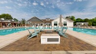 Located at 310 Northbend Drive in Charlotte, NC, this two-story community amenity center is designed to deliver a resort-caliber outdoor and indoor experience. The exterior architecture blends board-and-batten white siding, gray lap siding, a stone accent chimney, and pitched rooflines, creating a refined yet welcoming aesthetic set against a lush, tree-lined backdrop.

At the heart of the space is a generously scaled aquatic area featuring multiple connected pool sections. One pool offers in-water lounge chairs for a true resort feel, while another expansive swimming pool provides ample room for recreation. Sleek white sculptural chaise lounges line the sun shelf, and rows of traditional reclining chaise lounges are arranged along the broad tiled pool deck. A dedicated fire pit seating area, surrounded by Adirondack chairs, serves as a central gathering point on the paver terrace.

The pool deck is further outfitted with a series of private shaded cabanas, each furnished for relaxed lounging. A trellis-covered pergola zone adds additional shaded seating options. The surrounding landscape features manicured greenery and black wrought-iron fencing, enhancing privacy and visual appeal. Large windows on the clubhouse facade allow natural light to flow through the interior, seamlessly connecting indoor and outdoor amenity spaces throughout.