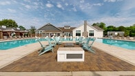 Located at 310 Northbend Drive in Charlotte, NC, this two-story community amenity center is designed to deliver a resort-caliber outdoor and indoor experience. The exterior architecture blends board-and-batten white siding, gray lap siding, a stone accent chimney, and pitched rooflines, creating a refined yet welcoming aesthetic set against a lush, tree-lined backdrop.

At the heart of the space is a generously scaled aquatic area featuring multiple connected pool sections. One pool offers in-water lounge chairs for a true resort feel, while another expansive swimming pool provides ample room for recreation. Sleek white sculptural chaise lounges line the sun shelf, and rows of traditional reclining chaise lounges are arranged along the broad tiled pool deck. A dedicated fire pit seating area, surrounded by Adirondack chairs, serves as a central gathering point on the paver terrace.

The pool deck is further outfitted with a series of private shaded cabanas, each furnished for relaxed lounging. A trellis-covered pergola zone adds additional shaded seating options. The surrounding landscape features manicured greenery and black wrought-iron fencing, enhancing privacy and visual appeal. Large windows on the clubhouse facade allow natural light to flow through the interior, seamlessly connecting indoor and outdoor amenity spaces throughout.