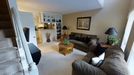 This two-floor residence offers a thoughtfully designed layout that balances comfort and functionality. The main living area is warm and inviting, featuring plush carpeting, neutral wall tones, and generous seating arranged around a classic round wooden coffee table. A built-in bookshelf unit serves as a natural divider between the living space and the adjoining areas, providing both storage and a decorative focal point. Framed artwork adorns the walls, adding a refined touch to the overall aesthetic. A floor lamp and carefully placed décor accents contribute to the cozy ambiance of the room. Adjacent to the living area, the kitchen is partially open to the main space via a raised countertop pass-through, enhancing the sense of connectivity throughout the home. The kitchen features stainless steel appliances and light cabinetry, creating a clean and functional cooking environment. Soft, recessed lighting throughout the main level ensures a welcoming atmosphere at any time of day. The two-floor configuration provides additional living or sleeping space, giving residents the benefit of distinct zones for relaxation and daily activities. Overall, this residence combines practical design with comfortable living in a well-maintained, move-in-ready setting.