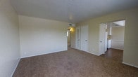 This single-story space presents a clean, functional layout centered around a generously sized main living area. The room is finished with wall-to-wall beige carpet, contributing to a warm and understated aesthetic. Smooth, pale yellow walls are complemented by white baseboards and trim, lending the space a bright, neutral character.

The textured ceiling adds a subtle visual element overhead, while a centrally located HVAC vent ensures climate control throughout. Natural light filters in from a window visible through the hallway, brightening the interior without overwhelming it.

Along the back wall, two white louvered-door closets provide practical storage solutions, while an additional door to the right offers access to what appears to be an adjoining room or bathroom, partially revealing white cabinetry. A wall-mounted light fixture in the hallway adds a functional lighting element.

The open flow between the main living space and the connecting hallway creates a sense of continuity throughout the unit. Overall, the space is characterized by its simple, well-maintained finishes and practical design, making it a versatile and livable environment.