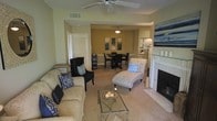 This single-story space showcases an open-concept layout that seamlessly connects the living and dining areas. The living room is anchored by a ceiling fan overhead and centers around a tiled fireplace with a white mantle, creating a welcoming focal point. Comfortable seating includes a neutral-toned sofa adorned with blue and white patterned throw pillows, a dark upholstered accent chair, and a tufted chaise lounge positioned near the fireplace. A glass-top coffee table with a decorative centerpiece ties the seating arrangement together. The flooring transitions from plush carpet in the living area to tile near the fireplace, adding visual definition to the space. An arched passageway leads into the dining area, which features a dark wood table with matching chairs and is lit by a classic pendant light fixture. Framed artwork in blue tones decorates the dining room wall, complementing the cohesive color palette throughout. Louvered closet doors and neutral beige walls contribute to the clean, understated aesthetic of this well-appointed space.