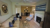 This single-story space showcases an open-concept layout that seamlessly connects the living and dining areas. The living room is anchored by a ceiling fan overhead and centers around a tiled fireplace with a white mantle, creating a welcoming focal point. Comfortable seating includes a neutral-toned sofa adorned with blue and white patterned throw pillows, a dark upholstered accent chair, and a tufted chaise lounge positioned near the fireplace. A glass-top coffee table with a decorative centerpiece ties the seating arrangement together. The flooring transitions from plush carpet in the living area to tile near the fireplace, adding visual definition to the space. An arched passageway leads into the dining area, which features a dark wood table with matching chairs and is lit by a classic pendant light fixture. Framed artwork in blue tones decorates the dining room wall, complementing the cohesive color palette throughout. Louvered closet doors and neutral beige walls contribute to the clean, understated aesthetic of this well-appointed space.