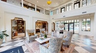 Located at 19020 Potts Park Ln in Davidson, NC, this beautifully appointed two-story clubhouse spans a generous footprint across two thoughtfully designed levels. The ground floor makes an immediate impression with its dramatic double-height great room, where soaring ceilings, large arched entry doors, and floor-to-ceiling windows flood the space with natural light. A striking black-and-white marble checkerboard floor anchors the main gathering area, furnished with elegantly patterned wingback chairs and a central seating arrangement. A grand curved staircase with dark wood treads and wrought iron railings serves as a stunning architectural centerpiece. Adjacent spaces include a dining area, a cozy sitting room framed by blue-painted millwork, and a charming office nook with bold plaid wallcovering. Upstairs, the second level opens to an expansive game room featuring rich herringbone hardwood floors, wood-paneled walls, a statement marble fireplace, and a full billiards table beneath a crystal chandelier. A built-in banquette nook with custom storage adds a playful, welcoming touch. The upper level also offers access to a covered outdoor terrace with herringbone brick pavers, ceiling fans, and open views of the surrounding Davidson community — ideal for relaxed outdoor gatherings.