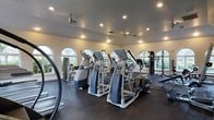 This fitness center offers a thoughtfully designed workout environment within a single-floor layout. The space is lined with a series of arched windows along multiple walls, flooding the interior with natural light and providing pleasant outdoor views of surrounding greenery, creating an inviting atmosphere for exercise. The high ceiling is fitted with recessed lighting and ceiling fans, ensuring consistent airflow and visibility throughout the room. The flooring is a durable dark rubber material well-suited for high-traffic fitness use. The cardio area is prominently featured, housing multiple elliptical trainers and stair-climber machines arranged in orderly rows, all equipped with individual display consoles. Along the perimeter, additional strength and cable-based equipment is visible, including a multi-station cable machine. Large mirrors on the walls further enhance the sense of openness and allow users to monitor their form during workouts. The overall ambiance is bright, clean, and functional, with the combination of natural light, modern equipment, and ample floor space contributing to a professional and welcoming fitness environment.