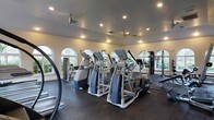 This fitness center offers a thoughtfully designed workout environment within a single-floor layout. The space is lined with a series of arched windows along multiple walls, flooding the interior with natural light and providing pleasant outdoor views of surrounding greenery, creating an inviting atmosphere for exercise. The high ceiling is fitted with recessed lighting and ceiling fans, ensuring consistent airflow and visibility throughout the room. The flooring is a durable dark rubber material well-suited for high-traffic fitness use. The cardio area is prominently featured, housing multiple elliptical trainers and stair-climber machines arranged in orderly rows, all equipped with individual display consoles. Along the perimeter, additional strength and cable-based equipment is visible, including a multi-station cable machine. Large mirrors on the walls further enhance the sense of openness and allow users to monitor their form during workouts. The overall ambiance is bright, clean, and functional, with the combination of natural light, modern equipment, and ample floor space contributing to a professional and welcoming fitness environment.