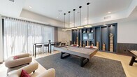 Located at 8555 East Raintree Drive in Scottsdale, Arizona, this thoughtfully designed amenity space spans a single floor and is divided into two distinct areas, each with its own character and purpose. The billiards lounge is bright and contemporary, anchored by a full-size pool table set atop a textured area rug over light hardwood flooring. A cluster of modern disc pendant lights hangs overhead, complementing the recessed ceiling lighting. A bold navy built-in shelving unit lines one wall, housing a flat-screen TV, curated books, and decorative accents. Comfortable lounge chairs and a high-top bar table with seating round out the space, while floor-to-ceiling sheer curtains diffuse ample natural light from large windows. A shuffleboard table and patterned accent wallpaper add further dimension to this zone. The adjoining lounge area offers a dramatically different atmosphere — moody and intimate, with deep burgundy walls, swan-motif wallpaper, and dark cabinetry. Curved caramel leather sofas and a marble-top coffee table create a welcoming seating arrangement, while gray upholstered booth seating backed by warm wood slat panels provides a restaurant-style social setting. A wall-mounted TV and integrated shelving with sconce lighting complete this refined, club-like retreat.