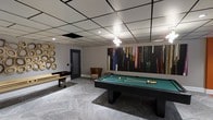 This well-appointed game room offers a dynamic and inviting atmosphere designed for social entertainment. At its center sits a full-size billiards table with a classic green felt surface, complemented by a gold-finished shuffleboard table along the wall — providing options for friendly competition. The room is anchored by a striking large-scale abstract painting in vivid multicolored hues, serving as a bold focal point against the neutral gray walls. A decorative gold circular wall sculpture adds an artistic accent, reinforcing the space's contemporary aesthetic. Recessed lighting and a modern chandelier provide layered illumination, while a drop-panel ceiling with dark grid framing lends a structured, polished finish overhead. Patterned carpet tiles in soft gray tones define the floor and add texture to the room. A cozy accent chair in the corner offers a comfortable spot to relax between games. The overall design balances functionality with style, making this a versatile and visually engaging space suited for leisure and socializing.