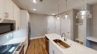 Located at 4930 Goose Creek Dr in Charlotte, NC, this single-floor residence offers a thoughtfully designed layout with contemporary finishes throughout. The kitchen serves as a central highlight, featuring shaker-style cabinetry in a soft neutral tone, a herringbone-patterned tile backsplash, stainless steel appliances including a refrigerator with ice dispenser, range, and built-in microwave. A center island with a white quartz countertop, undermount sink, and stylish glass pendant lighting provides both functionality and visual appeal. Warm hardwood-style flooring flows seamlessly through the kitchen and main living areas, lending a cohesive, modern feel. The open-concept living and dining space benefits from natural light through large windows with exterior views. Bedrooms are carpeted for comfort and include ceiling fans, with one bedroom featuring direct access to a walk-in closet. The primary bathroom is well-equipped with a white vanity, hardwood-style flooring, built-in shelving, and a walk-in closet accessible through a connecting doorway. Neutral warm-gray walls throughout the home create a calm, versatile backdrop. A built-in desk nook near the main living area adds a practical workspace. The overall design balances clean, modern aesthetics with functional, everyday living features.