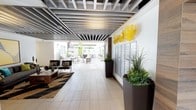 This single-floor space presents a thoughtfully designed interior that blends modern aesthetics with functional communal living. The lobby serves as the heart of the space, characterized by a striking exposed metal slat ceiling that adds an industrial yet refined touch to the overall design. Large-format light tile flooring runs throughout, lending a clean, cohesive look and enhancing the sense of openness.

A curated lounge area sits to one side, furnished with contemporary chairs, a wood coffee table, and a layered geometric area rug, creating an inviting spot to relax or socialize. Tall, dark planter boxes filled with lush greenery and sculptural plants are positioned throughout the corridor, introducing natural life and texture to the space.

A wall-mounted resident mailbox installation is neatly integrated along one side, accompanied by vibrant yellow glass wall art that adds a bold artistic focal point. The corridor opens seamlessly toward an outdoor courtyard, visible in the background, where umbrella-shaded seating arrangements and landscaping invite residents to enjoy the outdoors. Abundant natural light filters in from the courtyard, brightening the interior and creating a smooth indoor-outdoor flow that defines the ambiance of this well-appointed communal space.