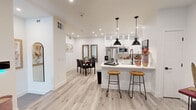 This thoughtfully designed single-floor residence showcases a clean, modern aesthetic anchored by light-toned hardwood flooring and crisp white walls throughout. The heart of the home is an open-concept living, dining, and kitchen area that flows seamlessly together. The chef's kitchen is a standout feature, outfitted with white shaker cabinetry extending to the ceiling with glass-front upper cabinets, quartz countertops with a striking veined pattern, a subway tile backsplash, stainless steel appliances including a range with a wall-mounted hood, and a large kitchen island with bar seating illuminated by a pair of bold matte black pendant lights. The adjoining living area offers a comfortable gathering space with recessed lighting and a modern ceiling fan. A dedicated dining area provides additional space for meals and entertaining. The bedroom is a serene retreat featuring plush carpeting, multiple windows with plantation shutters and elegant draping, and a stylish ceiling fan. The full bathroom is finished in neutral tones with tile flooring, a white vanity with a quartz countertop, matte black hardware and fixtures, and a tub-shower combination. Completing the layout is a dedicated laundry room equipped with a full-size washer and dryer set and clean tile flooring.