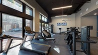 This fitness center is thoughtfully designed to accommodate a wide range of workout styles within a single, open-plan space. The facility is defined by soaring ceilings with a dark painted finish, exposed linear LED lighting, and a large ceiling fan that contribute to a sleek, contemporary aesthetic. Floor-to-ceiling windows along one wall flood the cardio zone with abundant natural light and offer views of the surrounding outdoors, creating an energizing atmosphere.

The cardio area features Matrix treadmills, elliptical trainers, and stair climbers, all positioned to take advantage of the natural light. Nearby, a dedicated cycling zone houses multiple stationary bikes equipped with individual screens, with dual wall-mounted televisions overhead for additional entertainment. A multi-functional cable and pulley strength rig anchors the strength training section, complemented by Matrix selectorized machines and a robust free weight area stocked with dumbbells, kettlebells, resistance bands, medicine balls, and a plyo box.

Large mirrors line the walls of the strength zone, visually expanding the space. A water fountain is conveniently located within the facility. Durable rubber flooring throughout provides safety and support, while recessed and track lighting in the secondary strength area ensures consistent illumination across all workout zones.