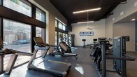 This fitness center is thoughtfully designed to accommodate a wide range of workout styles within a single, open-plan space. The facility is defined by soaring ceilings with a dark painted finish, exposed linear LED lighting, and a large ceiling fan that contribute to a sleek, contemporary aesthetic. Floor-to-ceiling windows along one wall flood the cardio zone with abundant natural light and offer views of the surrounding outdoors, creating an energizing atmosphere.

The cardio area features Matrix treadmills, elliptical trainers, and stair climbers, all positioned to take advantage of the natural light. Nearby, a dedicated cycling zone houses multiple stationary bikes equipped with individual screens, with dual wall-mounted televisions overhead for additional entertainment. A multi-functional cable and pulley strength rig anchors the strength training section, complemented by Matrix selectorized machines and a robust free weight area stocked with dumbbells, kettlebells, resistance bands, medicine balls, and a plyo box.

Large mirrors line the walls of the strength zone, visually expanding the space. A water fountain is conveniently located within the facility. Durable rubber flooring throughout provides safety and support, while recessed and track lighting in the secondary strength area ensures consistent illumination across all workout zones.