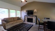 This single-floor residence offers a warm and inviting atmosphere anchored by a well-appointed living room. The space immediately draws attention to its vaulted ceiling, which adds a sense of openness and airiness to the room. A ceiling fan is centrally mounted, providing both function and a casual charm. The focal point of the living area is a classic brick fireplace, framed by a dark wood mantel that serves as a natural entertainment center, complete with a mounted flat-screen television and flanking table lamps that cast a soft, ambient glow. The walls are finished in a rich, earthy taupe tone that complements the warm brick and neutral carpeted flooring. A bold patterned area rug in deep navy and cream grounds the seating area, which features a comfortable tan sofa accented with decorative pillows. Natural light filters into the room through a window overlooking a lush outdoor tree line, adding a refreshing connection to the outdoors. The overall ambiance is cozy and functional, blending traditional design elements with comfortable, everyday living in a thoughtfully arranged single-story layout.