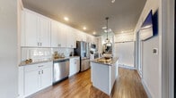 Located at 955 North Orlando Avenue in Maitland, Florida, this model unit offers a compelling look at the finishes and design details available in the community. The kitchen is the centerpiece of the space, featuring crisp white shaker-style cabinetry, granite countertops with subtle veining, a classic subway tile backsplash, and a full suite of stainless steel appliances including a French door refrigerator, dishwasher, microwave, and range. A central island with a granite top and pendant lighting above adds both functionality and style. Warm hardwood-style flooring flows throughout, complementing the clean, contemporary aesthetic. A glass partition wall separates the kitchen area from a cozy dining nook, which is appointed with a built-in banquette, textured stone accent wall, dark tray ceiling, and stylish pendant lighting — creating an intimate yet open feel. An additional kitchenette area with lighter wood cabinetry and a walk-in glass-enclosed shower demonstrates the versatility of the unit configurations available. Recessed lighting throughout keeps the space bright and inviting, while the open layout maximizes the sense of scale. This model provides an informative and visually rich representation of the high-quality interiors offered at this Maitland address.