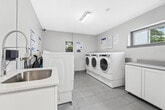 Located at 2360 Weston Rd in Toronto, Ontario, this shared laundry facility is a compact yet efficiently organized space designed to serve multiple users with ease. The room features a clean, modern aesthetic with crisp white walls and light grey tile flooring that runs throughout, contributing to a bright and hygienic atmosphere.

The laundry area is equipped with several front-loading washers and dryers arranged along both sides of the room, offering ample capacity for residents. The machines feature digital control panels, making operation straightforward and user-friendly. A dedicated utility area includes a white cabinetry-mounted stainless steel sink with a gooseneck faucet, ideal for hand-washing or pre-treating garments. A paper towel dispenser is conveniently mounted nearby.

Overhead fluorescent lighting ensures the space is well-lit throughout, while a small window on one wall allows for a degree of natural light and ventilation. Safety is addressed with a clearly visible fire extinguisher mounted near the entrance. Informational signage is posted on the walls to guide users on machine operation and available services. The solid grey entry door provides secure, controlled access to the facility. Overall, the space is practical, well-maintained, and thoughtfully arranged to accommodate everyday laundry needs efficiently.