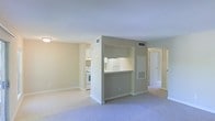 This single-floor space features an open, functional layout designed to maximize usability. The living area is finished with wall-to-wall carpeting in a neutral tone, providing a comfortable and cohesive feel. Tile flooring transitions into the kitchen zone, clearly delineating the two areas. The kitchen is partially separated by a half-wall partition, featuring a pass-through counter that connects it to the main living space. White cabinetry lines the kitchen walls, complemented by a white range and under-cabinet lighting that brightens the workspace. The neutral beige and cream color palette is consistent throughout, contributing to a clean, understated aesthetic. HVAC vents are integrated into both the walls and ceiling, ensuring climate control throughout. A hallway visible from the main living area leads to additional rooms, suggesting a practical and straightforward floor plan. The overall ambiance is understated and move-in ready, with simple finishes that provide a versatile backdrop suitable for a variety of interior styles.