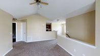 This single-floor home showcases a thoughtfully designed open-concept layout that maximizes both space and functionality. The main living area immediately impresses with its dramatic vaulted ceilings, which lend an expansive, airy quality to the room. A ceiling fan with a light fixture is centrally mounted, providing both comfort and illumination throughout the space. Soft, neutral beige walls and plush wall-to-wall carpeting create a warm and welcoming atmosphere throughout the living area. A pass-through window connects the living space to the kitchen, which features rich dark wood cabinetry and what appears to be stainless steel appliances, offering a practical yet stylish cooking space. A pendant light fixture adds a decorative touch above what is likely a dining or transitional area adjacent to the kitchen. Natural light filters in through windows, further brightening the interior. A staircase with a white-painted railing is visible to the right, suggesting access to additional levels or a lower entry. The floor plan flows seamlessly between spaces, making the home feel cohesive and well-connected. White trim, baseboards, and interior doors throughout provide a clean, classic finish that complements the neutral color palette.
