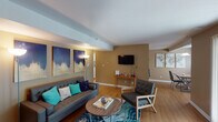 This single-floor residence offers a thoughtfully designed layout that balances comfort and modern style. The living room serves as the heart of the home, featuring warm hardwood-style flooring, a textured ceiling, and neutral tan walls accented by a structural beam detail. A gallery-style arrangement of bold, navy-and-gold wall art adds visual depth, while a wall-mounted flat-screen TV and floor lamps provide both function and ambiance. The seating area centers around a tufted gray sofa, a round wooden coffee table, and a vibrant blue marbled area rug that ties the space together.

The galley kitchen is efficiently designed with white cabinetry, dark granite-style countertops, stainless steel appliances including a refrigerator, range, microwave, and dishwasher, and hardwood-style flooring that continues from the main living area.

The primary bedroom is generously sized, featuring a large window with teal curtains that allow natural light, a tufted headboard, coordinating nightstands, and curated wall art. A second bedroom offers a comfortable layout with a window and ample natural light. An additional room visible in the floor plan appears to function as a home office or den.

The bathroom features clean white walls, hardwood-style flooring, and a shower-tub combination with a bold geometric-patterned curtain.