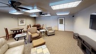 The game room is a generously sized, single-floor communal space designed to accommodate a variety of leisure and social activities. The room is finished with patterned carpeting throughout most of the floor, complemented by a tiled accent area near the main seating zone. Overhead, large recessed panel light fixtures provide consistent, even illumination across the space, supported by a ceiling fan that aids in air circulation.

The layout is thoughtfully divided into distinct activity zones. A lounge area features upholstered armchairs, an ottoman, and a mosaic-tiled coffee table arranged around a wall-mounted flat-screen television set atop a dark wood media console with storage. This media corner serves as a central entertainment hub. Nearby, a separate card or game table area offers a round wooden dining-style table with mission-style chairs, ideal for group activities or casual dining. Additional folding tables provide flexible space for various uses.

Decorative touches include framed artwork on the neutral-toned walls and a large faux indoor tree that adds warmth and a natural element to the room. A window along one wall allows natural light to filter in. The overall ambiance is comfortable, functional, and welcoming.