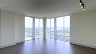 This single-floor space is defined by its striking floor-to-ceiling glass curtain wall that wraps around two exterior walls, flooding the interior with abundant natural light and offering expansive panoramic views of the surrounding urban landscape, including city buildings, greenery, and infrastructure below — indicative of a high-rise elevation. The open-plan layout feels generously proportioned, with clean white walls and smooth ceilings that complement the minimalist architectural design. A cylindrical structural column serves as a subtle architectural focal point near the center of the space, adding a modern design element while maintaining the openness of the floor plan. The rich, dark-toned hardwood flooring runs throughout, providing a warm contrast to the bright, airy atmosphere created by the surrounding glass. The corner window configuration maximizes both light penetration and sightlines, creating a seamless visual connection between the interior and the cityscape beyond. Overall, the space presents a clean, contemporary aesthetic with an emphasis on openness, natural light, and elevated urban views.