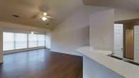 This single-story space showcases an open-concept layout that seamlessly connects the kitchen and living areas. The vaulted ceiling adds a sense of height and airiness, enhancing the overall spaciousness of the interior. Two ceiling fans are positioned throughout the space, providing both functional ventilation and ambient lighting.

The flooring throughout consists of rich, dark hardwood-style planks that add warmth and a polished aesthetic to the space. A white kitchen peninsula extends into the open area, creating a natural division between the kitchen and living zones while maintaining visual continuity. The countertop surface is clean and bright, complementing the neutral white walls.

Large windows along one wall allow generous natural light to filter into the living area, brightening the space and providing views of the surrounding exterior. The walls are finished in a crisp, neutral white tone, offering a versatile backdrop. The open layout, combined with the vaulted ceiling and natural light, creates an inviting and functional environment suitable for a variety of uses.