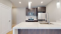 This single-floor space showcases a thoughtfully designed open-concept layout anchored by light hardwood flooring that flows throughout. The kitchen serves as a central highlight, featuring dark wood cabinetry with a rich grain finish, a dramatic waterfall-edge quartz island with an integrated sink, and a full suite of stainless steel appliances including a range, over-the-range microwave, and French door refrigerator. A marble-look backsplash ties the kitchen together, while cylindrical glass pendant lights add a refined touch overhead.

The open living area is generously proportioned, offering a clean, neutral palette with recessed lighting and a striking accent wall in muted teal that frames a sleek, modern linear fireplace. A large window invites natural light into the space. A separate room, likely a bedroom, features light hardwood floors, a ceiling fan with a brass finish, and a sliding glass door leading to an outdoor balcony with green views beyond.

The bathroom is elegantly appointed with large-format white tile flooring, a frameless glass walk-in shower clad in marble-look tile, a warm wood floating vanity with a marble countertop, and a wall-mounted toilet, creating a spa-like atmosphere throughout.