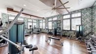 Located at 12 Digital Way in Maynard, MA, this fitness center offers a thoughtfully designed workout space that balances functionality with a vibrant aesthetic. The single-floor facility is characterized by rich hardwood flooring that runs throughout, creating a warm and polished foundation for the workout environment.

Distinctive tropical-print wallpaper in shades of green and white covers the accent walls, lending an energizing and resort-like ambiance to the space. Large, multi-pane windows line the walls, flooding the interior with abundant natural light and offering views of the surrounding exterior. Exposed dark wood ceiling beams add architectural character, while oversized ceiling fans with a sleek, modern design promote air circulation throughout.

The fitness center is comprehensively equipped to support a full range of training needs. Cardio equipment includes treadmills, stationary bikes, and elliptical machines, primarily positioned along the windowed walls. A dedicated strength-training area features cable machines, a multi-station cable tower, adjustable weight benches, and a full dumbbell rack displayed along a mirrored wall. The mirrored sections further enhance the sense of openness, reflecting natural light and allowing users to monitor their form. Recessed ceiling lighting supplements the natural light, ensuring the space remains bright and well-illuminated at all times.
