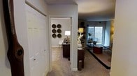 This single-story space is thoughtfully arranged, with a carpeted hallway serving as the central connector between distinct functional areas. To one side, a large white paneled closet provides ample built-in storage. A doorway off the hallway leads into a dedicated home office or study, where light-toned carpet, a patterned area rug, and a dark wood desk create a focused yet inviting workspace. A classic white floor lamp illuminates the room, while a set of six decorative woven baskets arranged in a grid pattern adds a striking artistic focal point to the walls.

The hallway opens into a broader living area, where the décor takes on a layered, eclectic character. Dark area rugs contrast the light carpet, and the room is furnished with a mix of dark wood and upholstered pieces. Potted plants introduce natural greenery, while shelving displays curated objects. A large window along the far wall draws in natural light and offers an outward view, brightening the space considerably. A textured ceiling runs throughout, and track lighting in the living area adds a modern, functional touch to the overall ambiance.