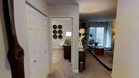 This single-story space is thoughtfully arranged, with a carpeted hallway serving as the central connector between distinct functional areas. To one side, a large white paneled closet provides ample built-in storage. A doorway off the hallway leads into a dedicated home office or study, where light-toned carpet, a patterned area rug, and a dark wood desk create a focused yet inviting workspace. A classic white floor lamp illuminates the room, while a set of six decorative woven baskets arranged in a grid pattern adds a striking artistic focal point to the walls.

The hallway opens into a broader living area, where the décor takes on a layered, eclectic character. Dark area rugs contrast the light carpet, and the room is furnished with a mix of dark wood and upholstered pieces. Potted plants introduce natural greenery, while shelving displays curated objects. A large window along the far wall draws in natural light and offers an outward view, brightening the space considerably. A textured ceiling runs throughout, and track lighting in the living area adds a modern, functional touch to the overall ambiance.