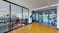 This contemporary fitness center is thoughtfully designed to accommodate a full range of workout activities across a single, expansive floor. The space is divided into distinct functional zones, seamlessly connected through sleek floor-to-ceiling glass partitions with black metal framing that lend the facility a polished, professional aesthetic.

The cardio and strength training area is outfitted with a comprehensive selection of equipment, including treadmills, ellipticals, stationary bikes, and various resistance machines. Large-scale black-and-white athletic murals adorn the walls, adding an energizing visual element to the workout environment. Linear LED lighting runs throughout the ceiling, ensuring bright, even illumination across the entire space.

An open studio zone features rich hardwood flooring and houses free weights, medicine balls, stability balls, and step platforms, making it well-suited for functional and group training. Floor-to-ceiling windows along this area flood the space with abundant natural light while offering expansive views of the surrounding outdoor landscape, including palm trees and the broader cityscape beyond.

A striking red spiral staircase serves as a design focal point near the glass-walled entrance, adding an architectural accent to the overall layout. The combination of modern materials, generous natural light, and a diverse equipment offering creates a motivating and professional fitness environment.