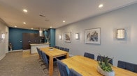This single-floor space offers a thoughtfully designed open-plan layout that seamlessly connects a spacious dining and meeting area with a fully equipped kitchen, making it well-suited for gatherings, events, or collaborative work sessions. The main room is anchored by two generous natural wood dining tables paired with upholstered navy blue cantilever chairs, providing ample seating for a large group. Soft gray carpeting runs throughout the main area, lending a refined, neutral backdrop that complements the bold teal accent walls. Natural light filters in through large windows along one wall, offering views of the surrounding streetscape. Recessed ceiling lighting and elegant wall sconces provide warm, layered illumination throughout. Framed artwork in neutral tones is displayed along the gray walls, adding a polished, curated aesthetic. The kitchen is a standout feature, boasting floor-to-ceiling dark wood cabinetry, a stainless steel island-mounted gas cooktop with a sleek overhead range hood, built-in wall ovens, and striking teal walls that create a bold, modern contrast. An adjacent room visible through a wide opening provides additional flexible space. The overall ambiance is contemporary and professional, balancing functionality with a clean, sophisticated design sensibility.