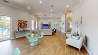 Located at 3305 Grant Cove Circle in Cape Coral, Florida, this single-story community space exudes a vibrant coastal charm throughout its well-appointed interior. The open-concept layout seamlessly connects the living, dining, and kitchen areas, creating a welcoming and functional environment.

The living area is generously proportioned, featuring warm hardwood-style flooring, soaring ceilings with crown molding, and an abundance of natural light streaming through multiple large windows with classic plantation shutters. The space is anchored by a bold entertainment wall with open wood-and-metal shelving and a large flat-screen TV, complemented by stylish seating arrangements that blend neutral tones with pops of coastal blue and teal.

The kitchen showcases an extended run of crisp white cabinetry topped with dark granite countertops, stainless steel appliances including a dishwasher, and wall-mounted screens — ideal for communal gatherings or presentations. A casual dining area nearby features playful aqua lattice chairs and a round wood table under elegant pendant lighting.

Two private office spaces provide a professional setting, each highlighted by coastal artwork, ornate drum pendant fixtures, and one featuring a dramatic deep-navy accent wall. Crown molding and coral-patterned area rugs tie the coastal theme throughout, making this a polished and inviting space.