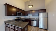 This single-floor residence offers a thoughtfully designed layout that balances comfort and functionality throughout. The kitchen serves as a standout feature, showcasing rich dark-wood cabinetry, granite countertops, and a full suite of stainless steel appliances including an electric range, built-in microwave, dishwasher, and refrigerator. A central island with matching granite provides additional prep space, while wood-look flooring and an elegant pendant light overhead complete the culinary space. The kitchen opens directly to a spacious living area finished with plush carpeting, creating a seamless and inviting flow between the two spaces. Soaring vaulted ceilings in the kitchen and living areas add a sense of openness and grandeur. The bedrooms are generously sized, featuring soft carpeting and windows that welcome natural light and views of the surrounding neighborhood. The bathroom is clean and functional, offering a tub-shower combination with a curved curtain rod, wood-look tile flooring, and a vanity with a granite countertop. Neutral tones throughout the residence create a bright, cohesive aesthetic that provides a versatile backdrop for a variety of interior styles.