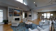 Located at 405 Thomas Burke Dr in Hillsborough, NC, this single-floor community space offers a thoughtfully designed environment that balances elegance with functionality. The main gathering area immediately impresses with its dramatic vaulted ceilings and clerestory windows that flood the room with natural light, creating an airy and open atmosphere. Neutral gray walls and warm hardwood flooring provide a refined backdrop throughout the space.

A classic white fireplace serves as a focal point in the lounge area, flanked by built-in shelving that adds both character and practicality. Comfortable upholstered seating in soft gray tones is arranged around a decorative wrought-iron coffee table atop a bold blue patterned area rug, lending the space a welcoming, curated feel. Large landscape artwork adorns the walls, adding warmth and visual interest.

Recessed lighting complements the natural light from the clerestory windows, ensuring the space is well-lit at any time of day. A long open corridor connects additional rooms toward the rear, suggesting versatile utility spaces or offices. A kitchenette or bar area is visible to one side, adding to the space's functionality. The overall ambiance is polished and inviting, making it well-suited for community gatherings, meetings, or relaxed socializing.