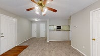 This single-story unit in Olive Branch, Mississippi, offers a functional and straightforward layout designed to maximize its comfortable footprint. The living area serves as the central hub of the space, finished with wall-to-wall carpet in a neutral beige tone and equipped with a ceiling fan with integrated lighting. Soft, neutral-gray walls throughout create a consistent, understated palette. A wood-toned entryway accent at the front door transitions into the carpeted main area, and a window allows natural light to filter in, offering views of surrounding greenery.

The kitchen is accessed via a pass-through counter with laminate countertops in a speckled brown finish. White cabinetry lines the walls above and below, providing ample storage, while appliances include an electric range and microwave. Wood-look flooring runs through the kitchen, distinguishing it from the carpeted living and bedroom areas.

The bedroom is a well-proportioned room with plush carpet, a ceiling fan, a window with blinds, a textured ceiling, and a closet. The bathroom features a full tub-shower combination with a curtain rod, white cabinetry above the toilet for additional storage, and wood-look flooring.