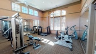 This fitness center offers a spacious, well-appointed workout environment designed to support a full range of exercise routines. The room features notably high ceilings accented by decorative crown molding and warm, neutral-toned walls that create an inviting atmosphere. Large arched and rectangular windows along multiple walls allow abundant natural light to flood the space, complemented by ceiling fans that promote comfortable air circulation throughout.

The facility is equipped with a comprehensive selection of cardio machines, including treadmills, upright and recumbent stationary bikes, and elliptical trainers. A multi-station cable and weight machine anchors the strength training area, providing versatile resistance exercise options. Floor-to-ceiling mirrors line one full wall, enhancing the sense of openness while allowing users to monitor their form during workouts.

Patterned low-pile carpeting covers the floor throughout, providing both comfort and durability underfoot. Recessed ceiling lighting supplements the natural light, ensuring the space remains bright during all hours. Glass-paned entry doors add a refined architectural touch, connecting the fitness center to the surrounding facility. Overall, the space balances functionality with an upscale aesthetic, making it a practical and pleasant environment for daily exercise.