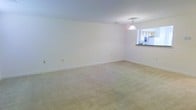 This two-floor space presents a clean, functional layout designed to maximize usability and flow. The main living area is generously sized, featuring wall-to-wall neutral-toned carpeting that adds warmth underfoot. Crisp white walls and standard ceilings create a bright, airy atmosphere throughout the room, while multiple electrical outlets are conveniently positioned along the baseboards.

A standout feature of the living area is its open connection to the kitchen via a pass-through counter with a dark countertop surface. This design element promotes an open feel between the two spaces while maintaining a visual distinction. A classic pendant light fixture hangs above the pass-through, providing focused illumination to the transitional zone. The kitchen, visible through the opening, features light-toned cabinetry and appears well-equipped.

The neutral color palette throughout — from the carpeting to the walls and ceiling — provides a versatile backdrop suitable for a variety of interior styles. The layout spans two floors, offering separation between living and private spaces, making it a practical and adaptable home.