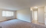 This single-floor residence offers a thoughtfully laid-out living space with a blend of comfort and functionality. The open living area is generously sized, featuring wall-to-wall carpeting, crisp white walls, and a large window fitted with horizontal blinds that allows natural light to fill the room. A baseboard heating unit ensures year-round comfort. The space transitions seamlessly into an additional carpeted room, which features a ceiling fan with integrated lighting, a bright window with blinds, and its own baseboard heating — ideal for use as a bedroom or flex space. Another carpeted room opens directly toward the entry, offering easy access to the light wood-tone flooring that extends through the transitional hallway areas. The kitchen is equipped with modern dark wood cabinetry, light stone-finish countertops, a stainless steel sink, a gas range with matching stainless steel over-the-range microwave, and a classic white subway tile backsplash. Light hardwood-style flooring completes the kitchen's clean, contemporary aesthetic. The bathroom is finished with white tile surrounding a full soaking tub and shower combination, a dark wood vanity with a white countertop, a brushed nickel faucet, a large mirror, and bright overhead lighting. Throughout the residence, white trim and neutral tones create a cohesive and inviting atmosphere.