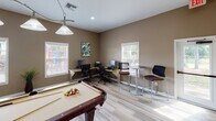 This single-floor amenity space is designed for both recreation and productivity, offering a well-appointed gathering area with a variety of functional zones. The main room features light wood-look flooring and warm taupe walls that create a welcoming, casual atmosphere throughout.

At the heart of the space sits a full-size pool table, serving as the focal point of the recreation area. A classic pendant light hangs overhead, while a wall-mounted flat-screen television provides an additional entertainment option. Seating includes bistro-style high-top tables with bar stools, as well as smaller game tables with chairs, offering flexible arrangements for socializing or gaming.

Natural light filters in through multiple windows along the walls, brightening the space and providing views of the surrounding greenery. A dedicated workstation area with computer setups and desk seating makes the space practical for those needing to work or study.

A standout feature is the wet bar area, finished with rich dark wood cabinetry, warm granite countertops, a stainless steel sink, and a dramatic floor-to-ceiling mosaic tile backsplash in earth tones. Three pendant lights hang above the counter, adding both function and style. Recessed lighting throughout complements the natural light, ensuring the space remains bright and inviting at all hours.