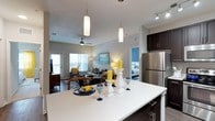This single-floor residence showcases a thoughtfully designed open-concept layout that seamlessly connects the kitchen, dining, and living areas. The kitchen is a standout feature, highlighted by a large white countertop island, dark espresso cabinetry, stainless steel appliances, and elegant pendant lighting that hangs above the island to create a warm, inviting atmosphere. Track lighting adds additional illumination throughout.

The living area flows naturally from the kitchen and is bathed in natural light courtesy of large windows that look out toward a balcony or outdoor space. A ceiling fan adds comfort to the airy environment, while the open sightlines create a generous sense of space throughout the common areas.

Neutral gray tones on the walls provide a clean and modern backdrop that complements the bold accent colors found in the furnishings and décor, including rich navy blues and vibrant yellows. A glimpse into the bedroom reveals a similarly well-lit and neatly appointed space, continuing the cohesive color palette.

The overall ambiance is contemporary and welcoming, with an emphasis on natural light, functional design, and stylish finishes that make this a comfortable and visually appealing living space.