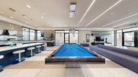 Located in Henderson, Nevada, this single-floor community space is a sophisticated lounge designed with a sleek, contemporary aesthetic. The open-concept layout seamlessly connects a fully equipped kitchen and an expansive living area, creating a cohesive and functional gathering environment.

The kitchen is a standout feature, showcasing floor-to-ceiling iridescent mosaic tile backsplash, dark cabinetry with polished hardware, and a large central island topped with bright white countertops and lined with modern bar stools. Pendant lighting and recessed fixtures illuminate the space, while a window frames views of the outdoors, flooding the area with natural light.

The living area is equally impressive, anchored by a stacked-stone fireplace that serves as a dramatic focal point. Large floor-to-ceiling windows and glass doors bathe the room in natural light while offering views of palm trees and outdoor amenities. Linear LED ceiling lighting adds a modern, architectural touch overhead. A full-size pool table takes center stage, complemented by plush blue sectional seating, tufted ottomans, and a wall-mounted entertainment unit. Large-format tile flooring in neutral tones runs throughout, tying the entire space together with a clean, polished finish.