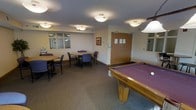 This single-floor amenity and recreation room offers a well-rounded space for both leisure and casual gatherings. The room is anchored by a full-sized pool table with a rich purple felt surface, providing a central focal point for entertainment. Adjacent to it, the seating area features multiple round tables surrounded by padded chairs, making it an ideal setting for group meetings or informal socializing. A separate windowed alcove adds architectural interest, offering a semi-enclosed nook that could serve as a quiet workspace or reading corner. The space is finished with neutral-toned walls and wall-to-wall carpeting in a muted grey, contributing to a warm and inviting atmosphere. Flush-mounted ceiling lights are evenly distributed throughout, ensuring consistent and practical illumination across the room. A framed piece of artwork adds a touch of character to the walls, while a set of double wooden doors provides a clearly defined entrance and exit. Overall, the room strikes a balance between functional utility and comfortable design, making it a welcoming communal space suited to a variety of activities.