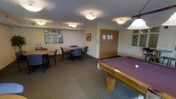 This single-floor amenity and recreation room offers a well-rounded space for both leisure and casual gatherings. The room is anchored by a full-sized pool table with a rich purple felt surface, providing a central focal point for entertainment. Adjacent to it, the seating area features multiple round tables surrounded by padded chairs, making it an ideal setting for group meetings or informal socializing. A separate windowed alcove adds architectural interest, offering a semi-enclosed nook that could serve as a quiet workspace or reading corner. The space is finished with neutral-toned walls and wall-to-wall carpeting in a muted grey, contributing to a warm and inviting atmosphere. Flush-mounted ceiling lights are evenly distributed throughout, ensuring consistent and practical illumination across the room. A framed piece of artwork adds a touch of character to the walls, while a set of double wooden doors provides a clearly defined entrance and exit. Overall, the room strikes a balance between functional utility and comfortable design, making it a welcoming communal space suited to a variety of activities.