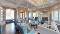 This sophisticated single-floor space makes a lasting impression through its refined architectural details and thoughtfully curated interiors. The entry area showcases dramatic arched columns, rich dark hardwood flooring, and crisp crown molding, setting a polished tone from the moment you arrive. Built-in shelving and teal accent seating add functional charm to the welcoming reception corridor, while frosted glass doors and large windows fill the space with natural light.

The centerpiece of the space is a breathtaking curved lounge room, where floor-to-ceiling wraparound windows offer sweeping outdoor views and bathe the interior in warm, natural light. A coffered tray ceiling with elegant crown molding frames a stunning orb chandelier that anchors the room. Plush neutral sofas, teal leather wingback chairs, and geometric accent tables create a comfortable yet upscale seating arrangement atop a layered area rug.

Adjacent to the curved lounge, a generously proportioned gathering area continues the refined aesthetic with additional tray ceilings, recessed lighting, arched doorways, and a classic white fireplace mantle. A decorative sunburst mirror and wall sconces add artistic flair, while the open sightlines between rooms enhance the sense of spaciousness. A kitchenette area is visible in the background, rounding out this beautifully appointed communal space.