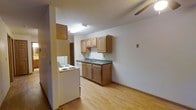 This single-floor unit offers a straightforward and functional layout designed for comfortable everyday living. The kitchen serves as a central feature of the space, outfitted with warm honey-toned wood cabinetry both above and below the countertops. Dark laminate countertops provide a subtle contrast to the light-colored cabinetry, and a stainless basin sink is positioned beneath upper cabinets for convenient use. White appliances, including a coil-top electric range and a full-sized refrigerator, are neatly integrated into the kitchen layout. Light hardwood-style flooring extends throughout, lending warmth and visual continuity to the space. The kitchen opens into an adjacent living area, creating an open, connected feel that maximizes the usable space. Overhead fluorescent lighting in the kitchen ensures the area is well-lit for cooking and daily tasks. The walls are finished in a clean, neutral white, providing a bright and airy atmosphere throughout. A doorway visible in the background hints at additional rooms, suggesting a practical floor plan designed to make efficient use of every square foot. Overall, the space presents a clean, move-in ready environment with classic, durable finishes suited for a wide range of occupants.