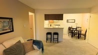 This single-floor residence offers an open-concept design that seamlessly integrates the living, kitchen, and dining areas into one cohesive space. The living area is anchored by plush wall-to-wall carpeting that adds warmth and comfort underfoot, providing a cozy setting for relaxation. A sofa with decorative pillows invites unwinding while remaining connected to the rest of the home. At the heart of the space, a peninsula-style kitchen counter with bar stool seating serves as a natural dividing element between the living and kitchen areas without sacrificing openness. The kitchen is equipped with dark espresso-toned upper cabinetry that provides a striking contrast against the light-colored walls and countertops. A full-size refrigerator and stovetop are neatly integrated into the kitchen layout. Adjacent to the kitchen, the dining area accommodates a table and chair set, making it ideal for both everyday meals and casual entertaining. A framed decorative piece adorns the dining area wall, adding a touch of personality. The soft, neutral color palette throughout — featuring warm beige and cream tones — creates a welcoming and harmonious ambiance. A doorway visible from the living area provides access to additional rooms, completing this well-organized and functional single-floor layout.