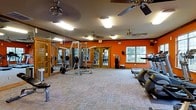 This fitness center is a fully appointed workout facility designed to support a comprehensive exercise routine. The space is finished with bold orange accent walls that energize the environment, complemented by neutral carpet tile flooring that spans the entire area. Multiple ceiling fans are evenly distributed overhead, ensuring consistent airflow and comfort during workouts.

The strength training area features a multi-station weight machine, a free-weight bench press setup, and an assortment of weight plates, providing a solid foundation for resistance training. Full-length mirrors line one wall, allowing users to monitor their form and enhancing the sense of space throughout the room.

On the cardio side, treadmills and other aerobic equipment are positioned near the windows, which provide natural light and views of the surrounding greenery outside. The windows bring in a pleasant connection to the outdoors without compromising the facility's climate control.

Access to the space is provided through elegant wood-framed glass doors, adding a warm architectural touch to the functional interior. Emergency exit signage and safety equipment are clearly visible, reflecting attention to occupant safety. Overall, this fitness center offers a well-organized, energetic, and practical environment suited for a wide range of exercise activities.