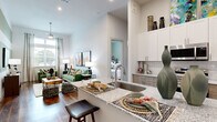 This single-story residence is defined by its open-concept design and contemporary finishes. The kitchen serves as a central hub, featuring a large granite-topped island with an undermount sink and bar seating, sleek taupe cabinetry, a subway tile backsplash, stainless steel appliances including a French door refrigerator, and a built-in dishwasher. A dedicated laundry area is conveniently tucked nearby. The layout flows seamlessly from the kitchen into the living area, which showcases soaring ceilings, rich wood-style flooring, and a bold accent wall with sage green horizontal stripes. Large windows flood the space with natural light, while a ceiling fan adds a functional touch. A sliding barn-style door adds architectural character along the corridor. The bedroom is a comfortable retreat with natural light from a side window, ample floor space, and access to a well-appointed bathroom featuring a soaking tub, granite vanity, and wood-style flooring. A dedicated walk-in closet with built-in shelving and a clothing rod provides generous storage. Throughout the residence, the combination of neutral white walls, warm wood tones, and modern fixtures creates a cohesive, inviting ambiance.